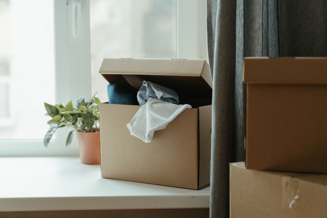Carton ouvert contenant des vêtements près d’une fenêtre, évoquant un déménagement ou une relocalisation.